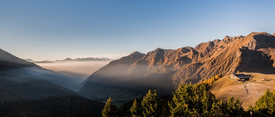 Le località dell’area vacanze Rio Pusteria L'immagine mostra un pittoresco paesaggio montano all'alba o al tramonto, con valli profonde coperte di nebbia e pendii di montagna illuminati dal sole che brillano in calde tonalità arancioni. Su una cresta montuosa a destra dell'immagine si trova un rifugio di montagna, circondato da colline erbose e alberi coniferi.