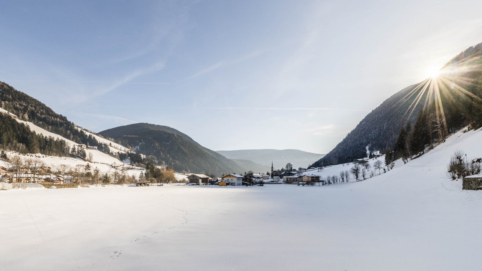 Quando si dice idilliaco… ecco a voi Fundres L'immagine mostra un paesaggio invernale innevato in una valle. In lontananza si vede un piccolo villaggio, circondato da colline e boschi coperti di neve. Il sole splende luminoso e irradia sopra la cresta della montagna a destra dell'immagine.