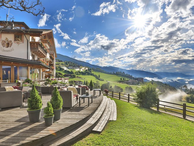 Benvenuti nell’Area Vacanze Sci e Malghe Rio Pusteria! Terrazza di un albergo con vista su paesaggio montano verde e sole splendente