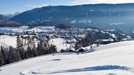 Voglia di slittino? Venite a Terento! L'immagine mostra un paesaggio montano innevato con un piccolo villaggio nella valle, circondato da colline e montagne. Il cielo è limpido e blu, e in primo piano si vedono tracce nella neve su un pendio dolce.