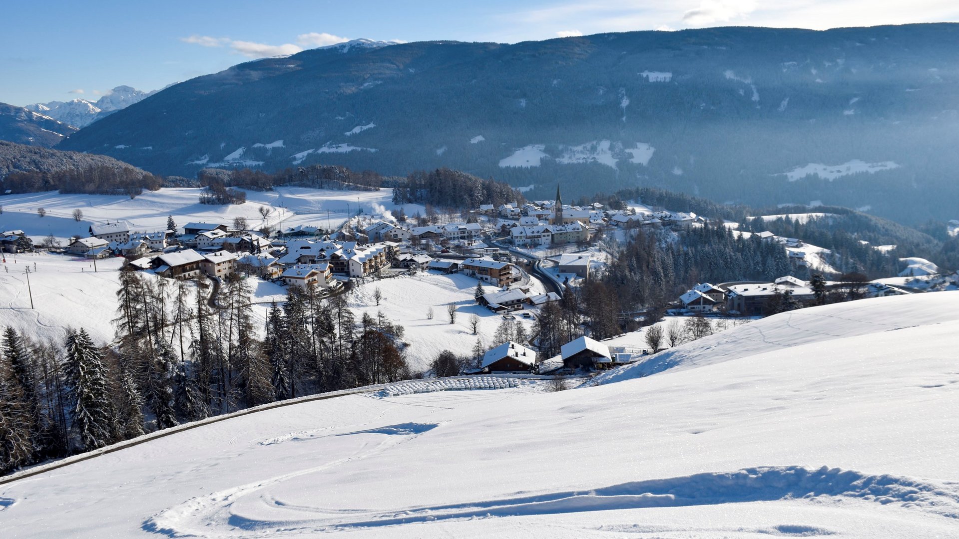 Voglia di slittino? Venite a Terento! L'immagine mostra un paesaggio montano innevato con un piccolo villaggio nella valle, circondato da colline e montagne. Il cielo è limpido e blu, e in primo piano si vedono tracce nella neve su un pendio dolce.