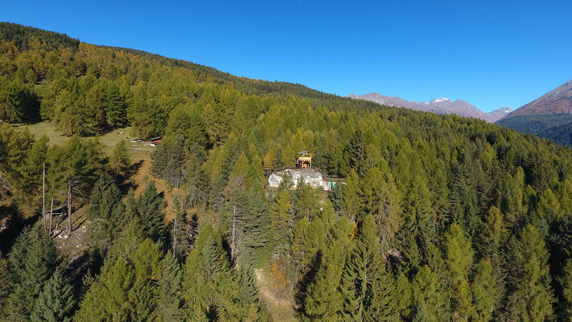 La zona ricreativa bunker di Spinga L'immagine mostra un piccolo edificio situato su una collina boscosa, circondato da una fitta foresta e montagne sullo sfondo. Il cielo è limpido e blu, e il terreno è coperto da molti alberi verdi.