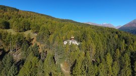 La zona ricreativa bunker di Spinga L'immagine mostra un piccolo edificio situato su una collina boscosa, circondato da una fitta foresta e montagne sullo sfondo. Il cielo è limpido e blu, e il terreno è coperto da molti alberi verdi.