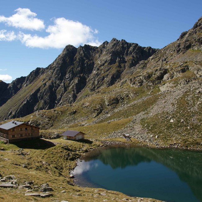 Tiefrastenhütte L'immagine mostra un rifugio di montagna accanto a un tranquillo e limpido lago alpino in un paesaggio roccioso alpino. Ripide montagne frastagliate si ergono sullo sfondo sotto un cielo blu con nuvole sparse.