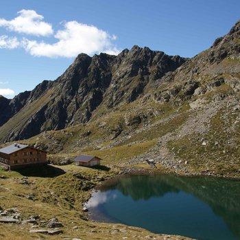 Sapori in malga L'immagine mostra un rifugio di montagna accanto a un tranquillo e limpido lago alpino in un paesaggio roccioso alpino. Ripide montagne frastagliate si ergono sullo sfondo sotto un cielo blu con nuvole sparse.