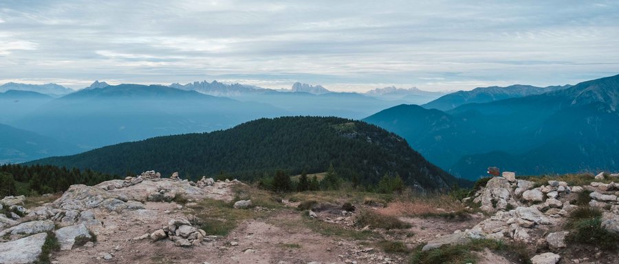 Le località dell’area vacanze Rio Pusteria L'immagine mostra un paesaggio montano con più livelli di colline e montagne che si estendono in lontananza. In primo piano c'è una superficie rocciosa, mentre sullo sfondo si vedono colline boschive e catene montuose più lontane sotto un cielo nuvoloso.