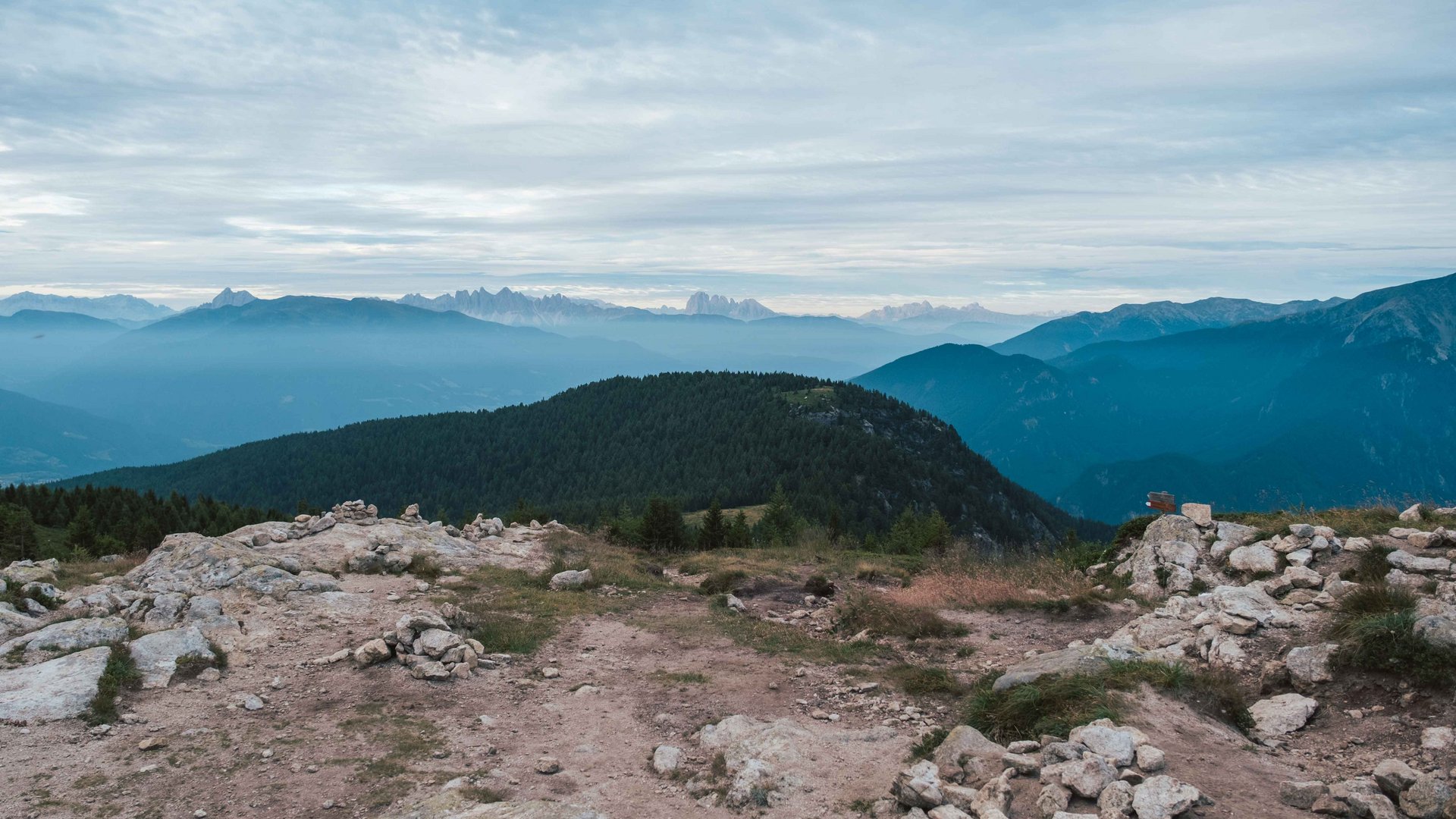 Valles - Jochtal - Steinermandl L'immagine mostra un paesaggio montano con più livelli di colline e montagne che si estendono in lontananza. In primo piano c'è una superficie rocciosa, mentre sullo sfondo si vedono colline boschive e catene montuose più lontane sotto un cielo nuvoloso.