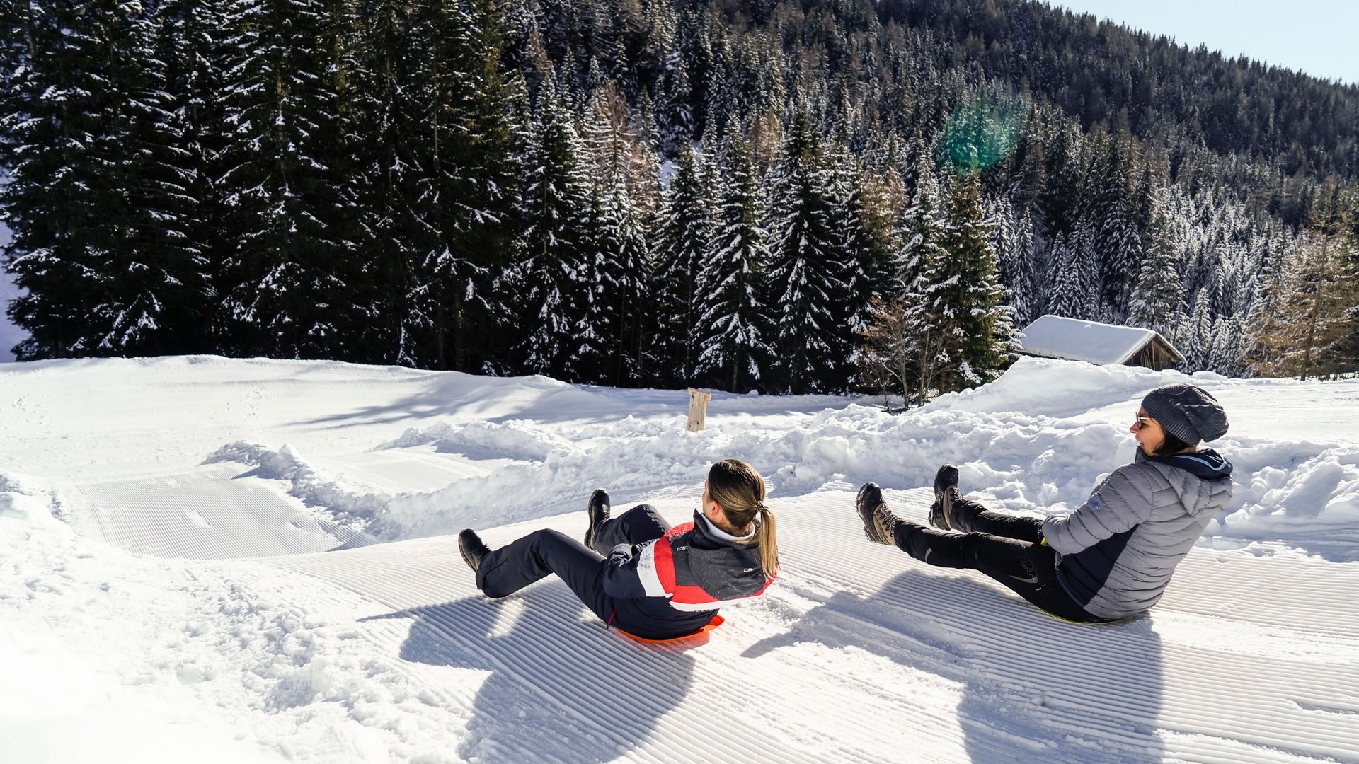 La Valle di Altafossa, presso Maranza L'immagine mostra due persone che scendono una pista da sci preparata su una slitta. Indossano abbigliamento invernale e sono circondate da alberi innevati e da una collina boscosa sullo sfondo. È una giornata chiara e soleggiata.