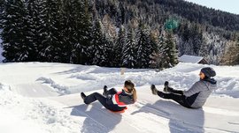 La Valle di Altafossa, presso Maranza L'immagine mostra due persone che scendono una pista da sci preparata su una slitta. Indossano abbigliamento invernale e sono circondate da alberi innevati e da una collina boscosa sullo sfondo. È una giornata chiara e soleggiata.