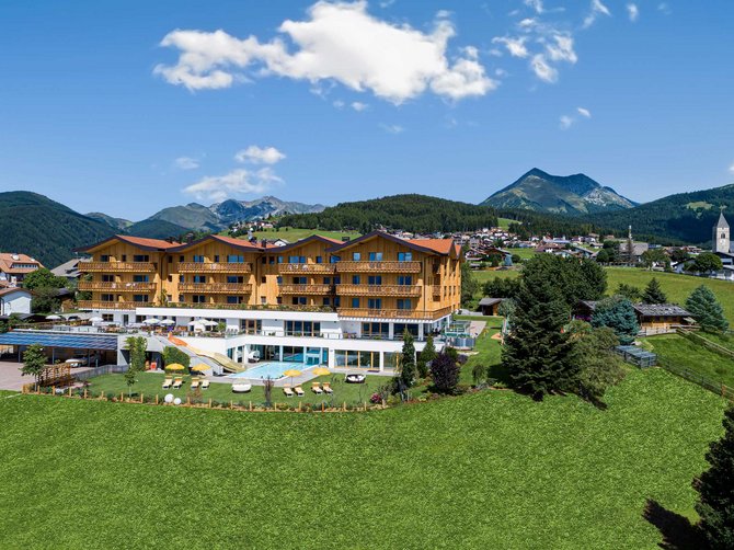Benvenuti nell’Area Vacanze Sci e Malghe Rio Pusteria! L'immagine mostra un grande edificio alberghiero moderno in un ambiente alpino. In primo piano c'è un prato verde, mentre sullo sfondo si vedono montagne e una piccola città.