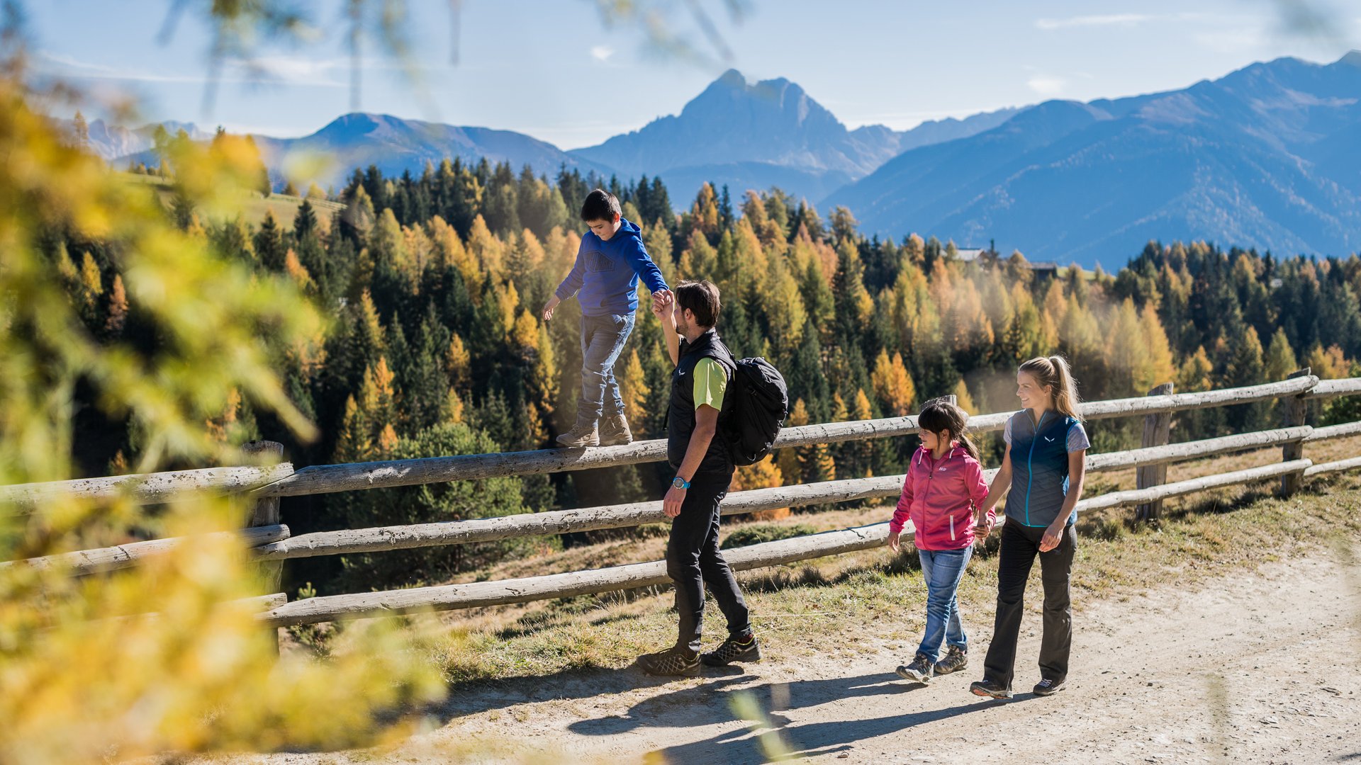 L’Alpe di Rodengo e Luson L'immagine mostra una famiglia che fa escursioni su un sentiero in montagna. Un bambino si equilibra su una recinzione di legno mentre il padre tiene la sua mano, e sullo sfondo si vedono alberi autunnali colorati e montagne.