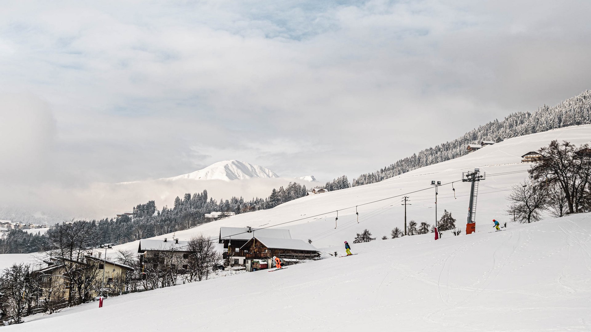 Vacanze sugli sci con i bambini a Terento L'immagine mostra un paesaggio innevato con una seggiovia e diversi sciatori su una pista. Sullo sfondo si vedono colline e montagne innevate, parzialmente coperte da nuvole.
