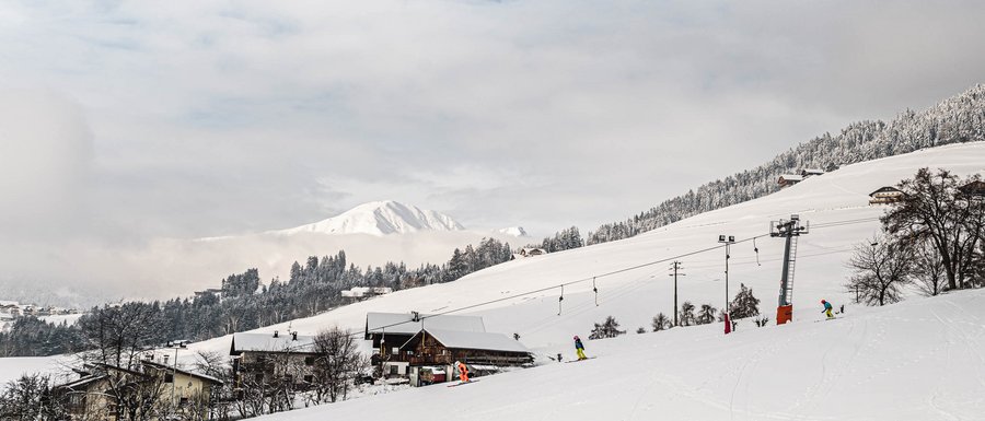 Le località dell’area vacanze Rio Pusteria L'immagine mostra un paesaggio innevato con una seggiovia e diversi sciatori su una pista. Sullo sfondo si vedono colline e montagne innevate, parzialmente coperte da nuvole.