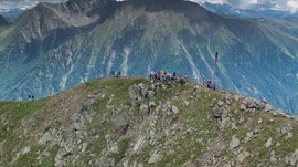 La piattaforma panoramica Gitschberga a Maranza (BZ) L'immagine mostra un gruppo di persone che stanno su una vetta rocciosa di montagna. Sullo sfondo si vedono impressionanti catene montuose verdi coperte da nuvole.