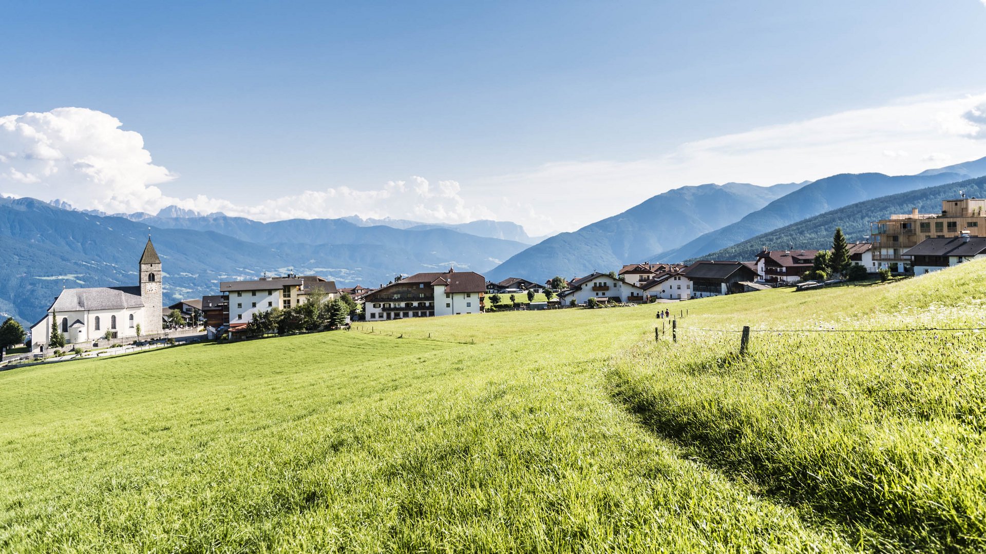 Conoscete già Maranza? L'immagine mostra un pittoresco villaggio con una chiesa sul lato sinistro, circondato da prati verdi e case sparse. Sullo sfondo si estendono montagne boscose sotto un cielo limpido e azzurro con poche nuvole.