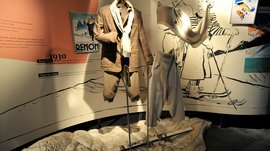 Il Mondo del Loden a Vandoies L'immagine mostra una mostra con abbigliamento da sci degli anni '30, presentato su manichini da vetrina. Sullo sfondo si vedono illustrazioni di sciatori e un vecchio poster che rappresentano la moda e lo sci di quell'epoca. Un paio di sci giacciono a terra davanti all'abbigliamento.