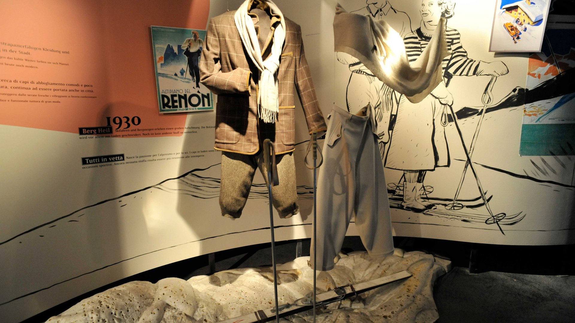 Il Mondo del Loden a Vandoies L'immagine mostra una mostra con abbigliamento da sci degli anni '30, presentato su manichini da vetrina. Sullo sfondo si vedono illustrazioni di sciatori e un vecchio poster che rappresentano la moda e lo sci di quell'epoca. Un paio di sci giacciono a terra davanti all'abbigliamento.