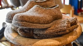 L’Avvento di Terento L'immagine mostra un paio di pantofole grigie in feltro che giacciono su uno sgabello. Sotto le pantofole c'è un maglione lavorato a maglia spesso e piegato, il tutto con un aspetto accogliente e fatto a mano.