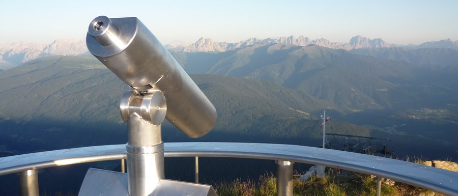 Le località dell’area vacanze Rio Pusteria L'immagine mostra un binocolo fissato permanentemente su un punto panoramico, rivolto verso un paesaggio montano ampio. Sullo sfondo si vedono catene montuose e valli sotto un cielo azzurro limpido.