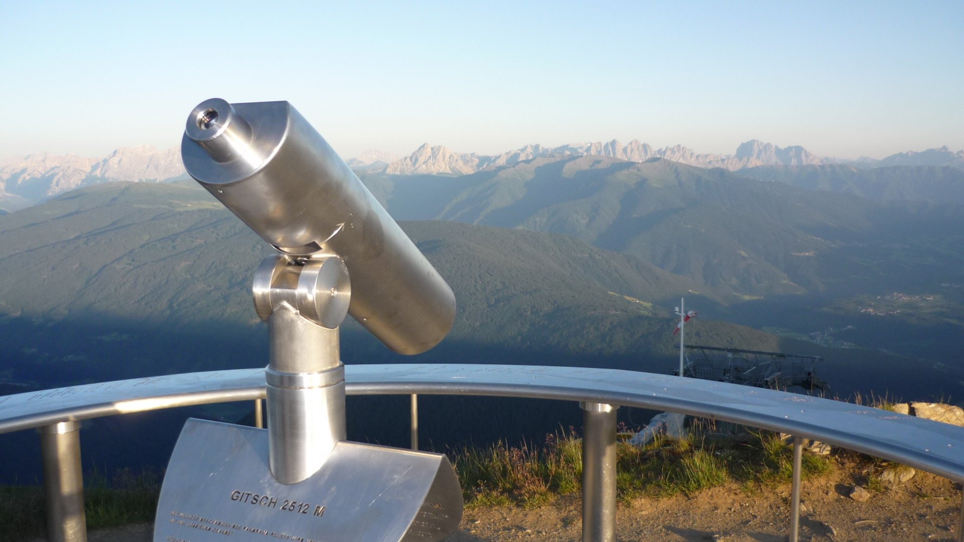 La piattaforma panoramica Gitschberga a Maranza (BZ) L'immagine mostra un binocolo fissato permanentemente su un punto panoramico, rivolto verso un paesaggio montano ampio. Sullo sfondo si vedono catene montuose e valli sotto un cielo azzurro limpido.
