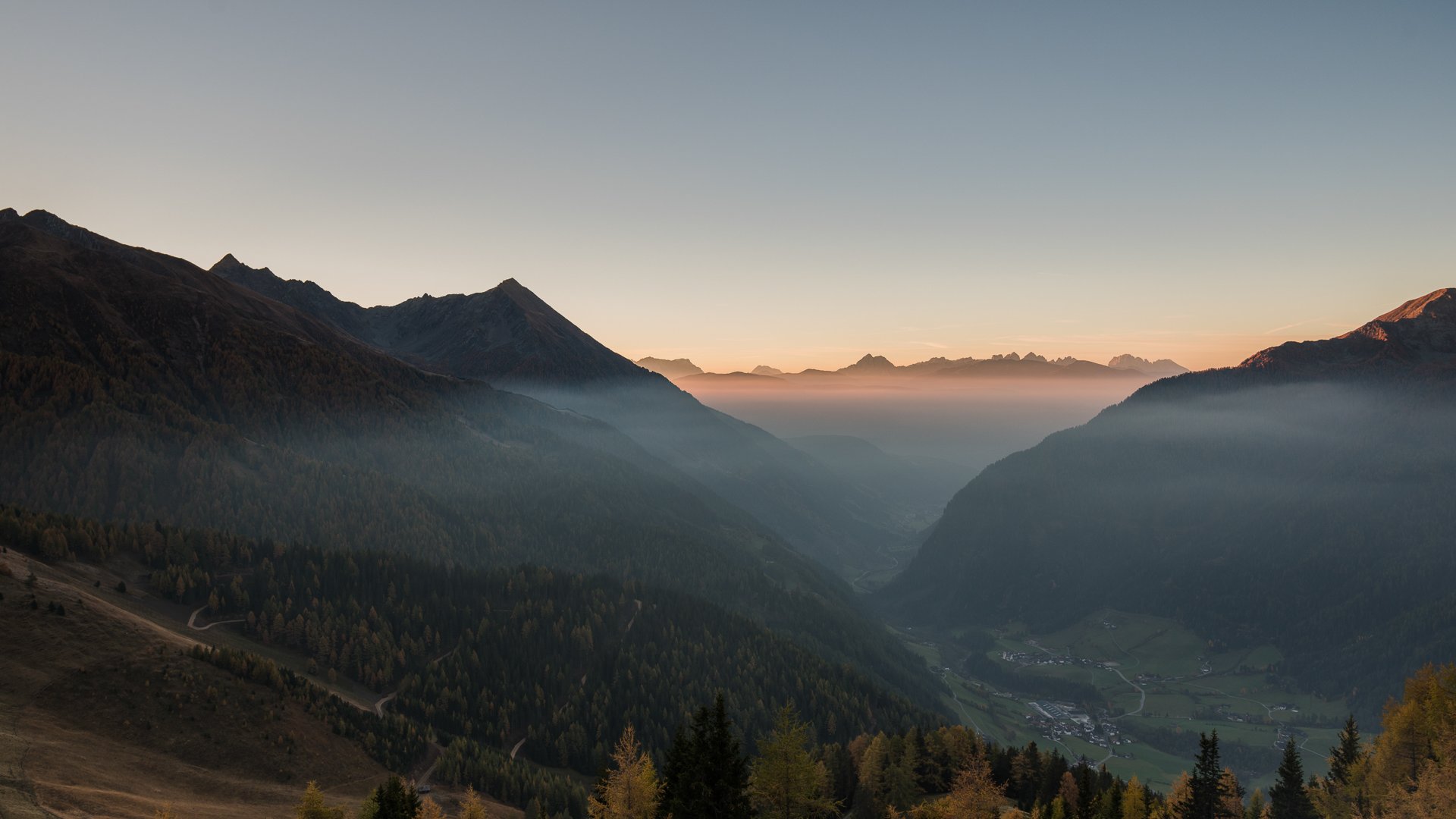 Quando si dice idilliaco… ecco a voi Fundres L'immagine mostra un vasto paesaggio montano durante l'alba o il tramonto. La nebbia riempie le valli tra le montagne, mentre le cime sono illuminate da una luce morbida e dorata, creando un'atmosfera pacifica e pittoresca.