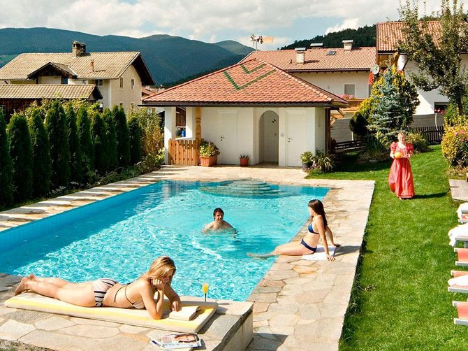 Benvenuti nell’Area Vacanze Sci e Malghe Rio Pusteria! L'immagine mostra una piscina all'aperto in un'area giardino ben curata, circondata da sdraio. Due donne prendono il sole vicino alla piscina, mentre una persona nuota nell'acqua. Sullo sfondo si vedono diversi edifici e montagne, che creano un ambiente tranquillo e idilliaco.