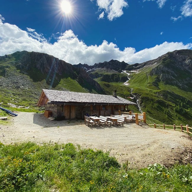 Kröllhütte-Eisbruggalm Una baita di montagna in legno e pietra si trova su un soleggiato e verde prato di montagna, circondata da alte montagne rocciose. Davanti alla baita ci sono diversi tavoli e panche di legno.