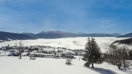 Conoscete già Maranza? L'immagine mostra un paesaggio innevato con un piccolo villaggio in primo piano, mentre sullo sfondo si estende un mare di nuvole. Sopra si ergono maestose montagne innevate sotto un cielo azzurro e limpido.