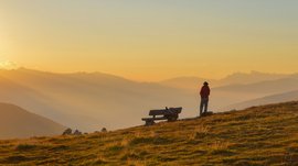 Valles - Jochtal - Steinermandl L'immagine mostra una persona che si trova in un prato di montagna ricoperto di erba e guarda lontano, mentre il sole tramonta all'orizzonte e illumina il cielo con calde tonalità di giallo. Accanto alla persona c'è una panchina di legno e sullo sfondo si vedono dolci catene montuose avvolte nella nebbia.