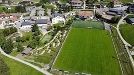Il Parco Active a Terento L'immagine mostra una veduta aerea di un villaggio con un grande prato verde che serve come campo da calcio. Intorno al campo ci sono diversi edifici, tra cui case con pannelli solari sui tetti, un campo da minigolf e sentieri che attraversano un paesaggio verde curato.