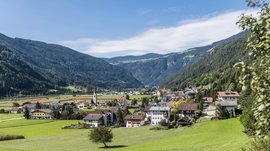 Vandoies: il cuore artigianale della Val Pusteria L'immagine mostra un paesaggio pittoresco con prati verdi e un villaggio nella valle. Sullo sfondo si vedono colline boscose e al centro del villaggio si erge una chiesa con una torre alta e appuntita.