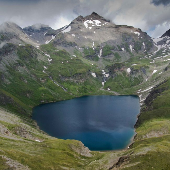Wilde Kreuzspitze L'immagine mostra un lago di montagna situato in alto, circondato da ripide pendici verdi e cime innevate. Il cielo è coperto di nuvole, il che conferisce alla scena un'atmosfera drammatica.