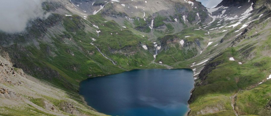 Wilde Kreuzspitze L'immagine mostra un lago di montagna situato in alto, circondato da ripide pendici verdi e cime innevate. Il cielo è coperto di nuvole, il che conferisce alla scena un'atmosfera drammatica.