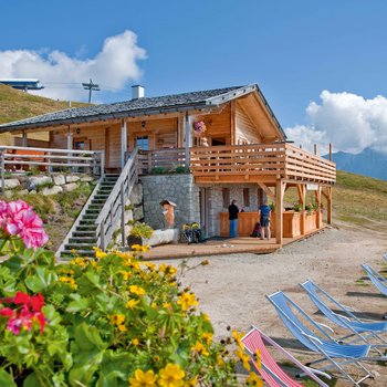 Sapori in malga L'immagine mostra una accogliente baita di montagna in legno con una grande terrazza e cassette di fiori. In primo piano ci sono fiori colorati e diverse sdraio. Persone stanno in piedi e chiacchierano davanti alla baita. Sullo sfondo ci sono colline verdi e montagne sotto un cielo azzurro con poche nuvole.