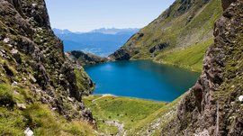 La Valle di Altafossa, presso Maranza L'immagine mostra un lago di montagna di un blu profondo, circondato da ripide pendici verdi. Sullo sfondo si vedono altre catene montuose e un cielo azzurro limpido, che trasmettono un paesaggio tranquillo e maestoso.