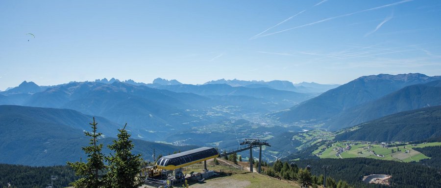 Le località dell’area vacanze Rio Pusteria L'immagine mostra un'ampia paesaggio montano sotto un cielo azzurro e limpido. In primo piano c'è una stazione della funivia sulla vetta di una montagna, mentre sullo sfondo si vedono diverse catene montuose e una valle con villaggi sparsi e boschi. Due parapendii volano in alto nel cielo sopra la scena.