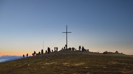 Valles - Jochtal - Steinermandl L'immagine mostra un gruppo di persone raccolte intorno a una grande croce di vetta su una cima. Il cielo è limpido e si presenta in delicati toni di blu e arancione, indicando l'alba o il tramonto.