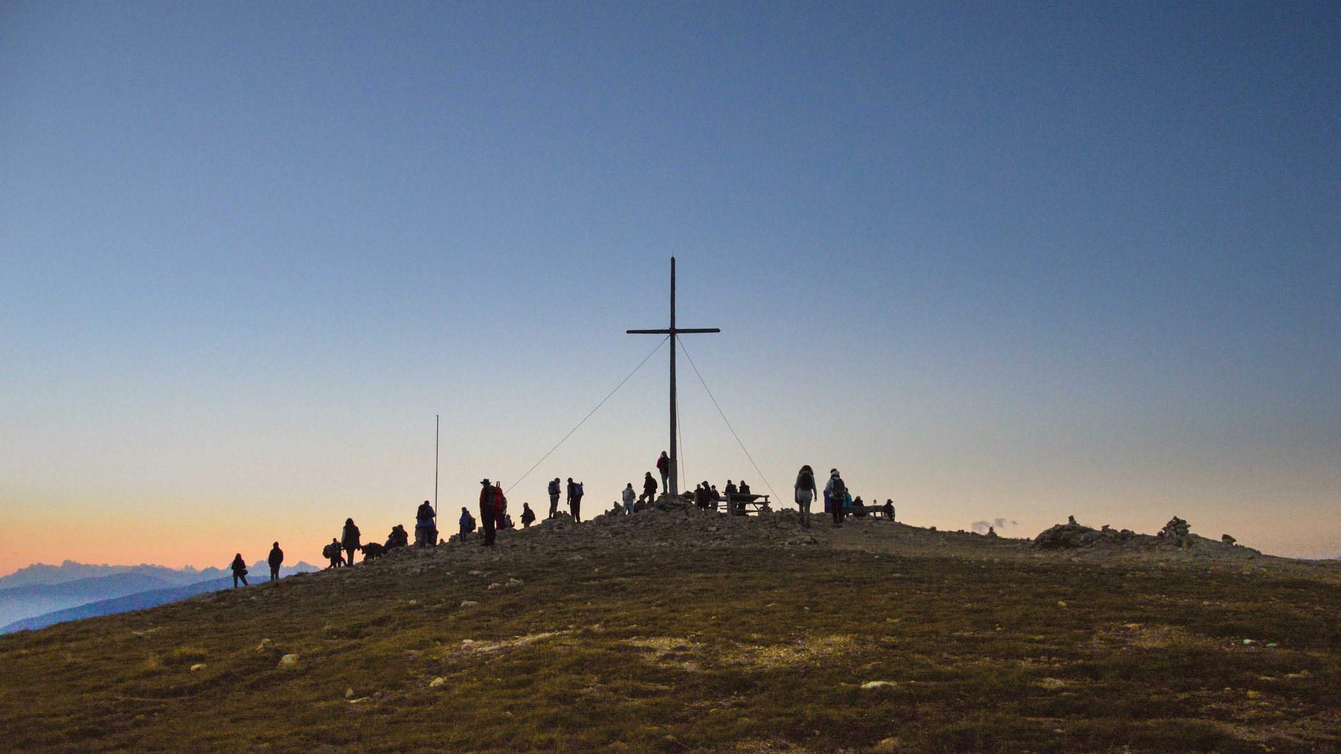Valles - Jochtal - Steinermandl L'immagine mostra un gruppo di persone raccolte intorno a una grande croce di vetta su una cima. Il cielo è limpido e si presenta in delicati toni di blu e arancione, indicando l'alba o il tramonto.