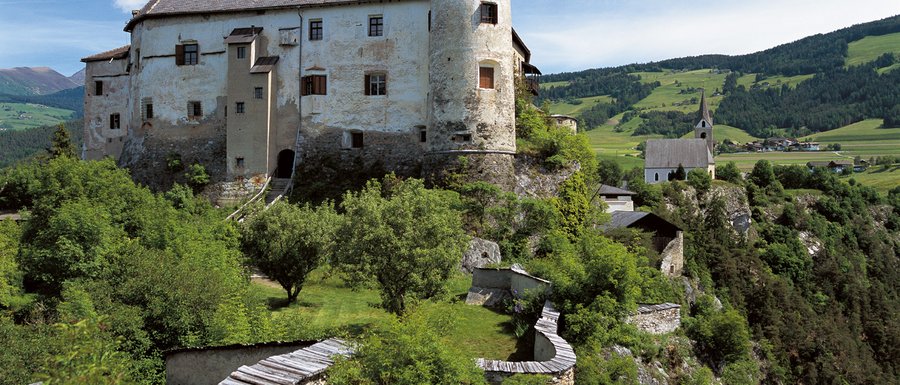 Le località dell’area vacanze Rio Pusteria L'immagine mostra un vecchio castello su una collina verde, circondato da alberi e da un muro di pietra. Sullo sfondo si vede una chiesa e un paesaggio rurale.