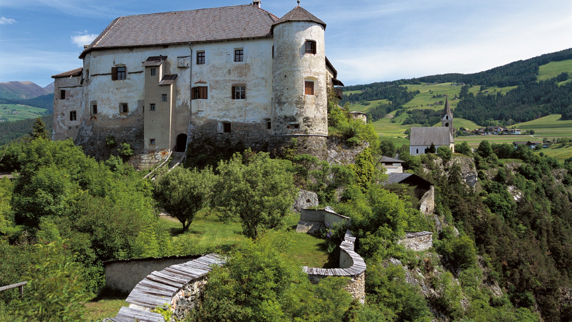 Alla scoperta di Castel Rodengo L'immagine mostra un vecchio castello su una collina verde, circondato da alberi e da un muro di pietra. Sullo sfondo si vede una chiesa e un paesaggio rurale.