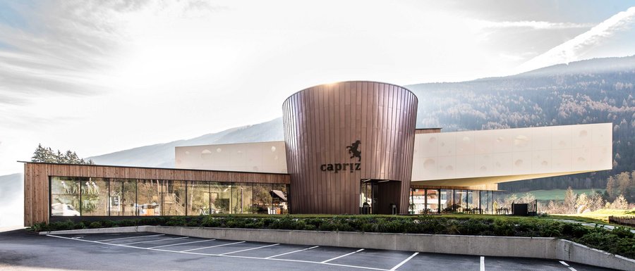 Le località dell’area vacanze Rio Pusteria L'immagine mostra un edificio moderno con una facciata rotonda color rame e grandi finestre di vetro. Sull'edificio è visibile la scritta "Capriz" e davanti c'è un parcheggio vuoto, mentre sullo sfondo si riconoscono colline e montagne boschive.