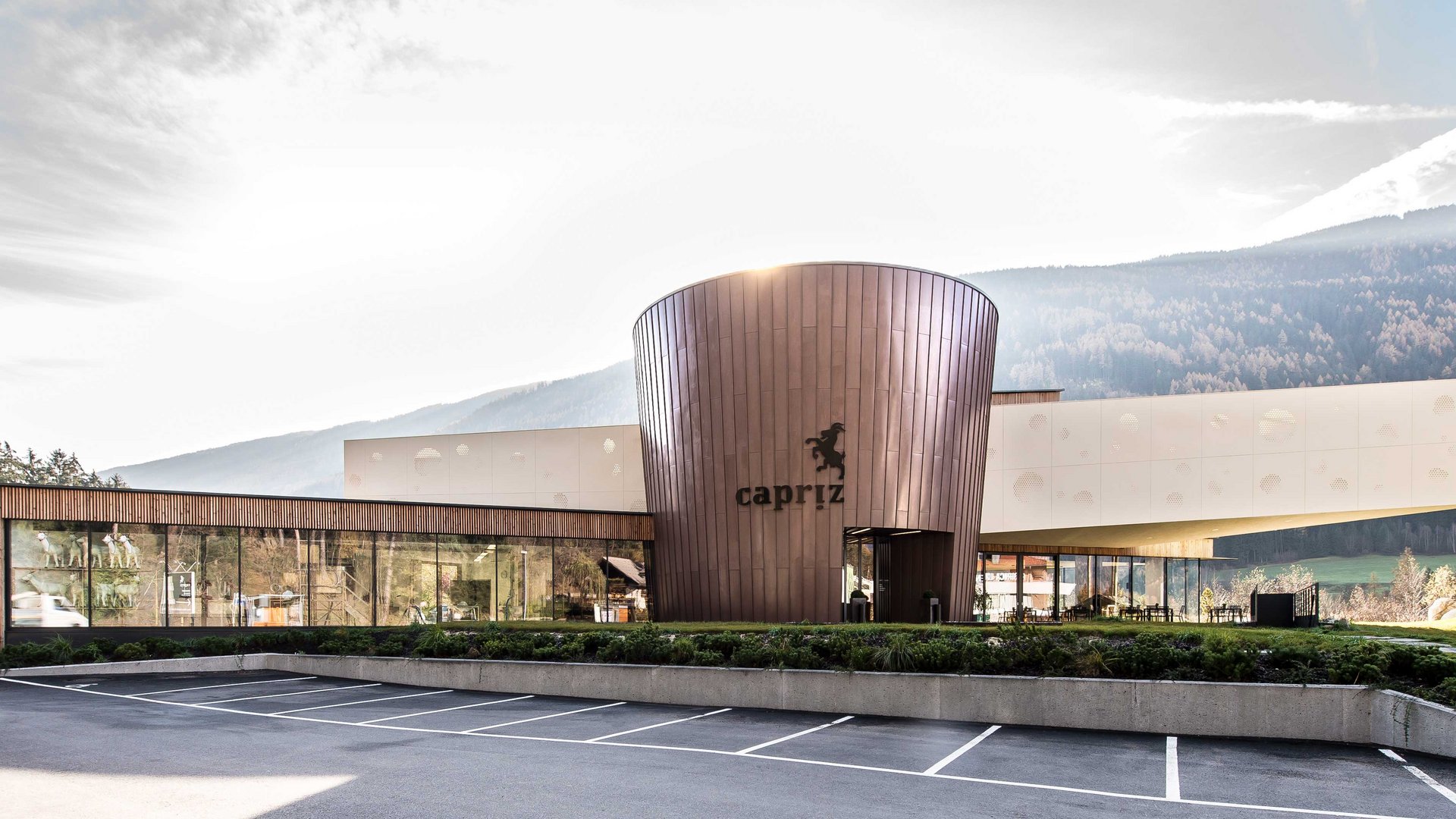 Il caseificio Capriz a Vandoies L'immagine mostra un edificio moderno con una facciata rotonda color rame e grandi finestre di vetro. Sull'edificio è visibile la scritta "Capriz" e davanti c'è un parcheggio vuoto, mentre sullo sfondo si riconoscono colline e montagne boschive.