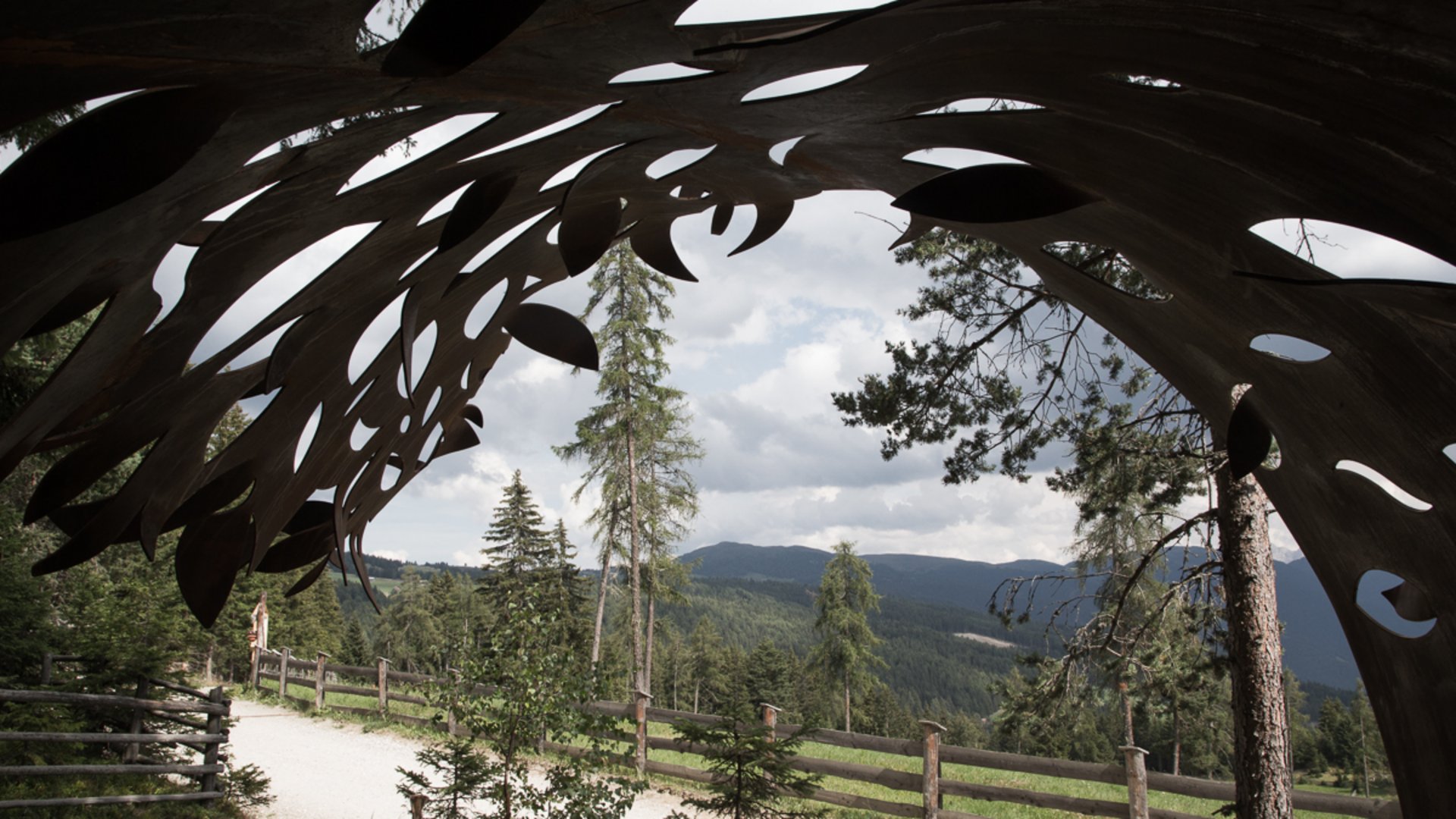 Il Sentiero della Creazione a Rodengo L'immagine mostra una vista attraverso una scultura metallica con forme organiche. Sullo sfondo si vedono alberi, un sentiero con una recinzione in legno e colline boschive sotto un cielo nuvoloso.