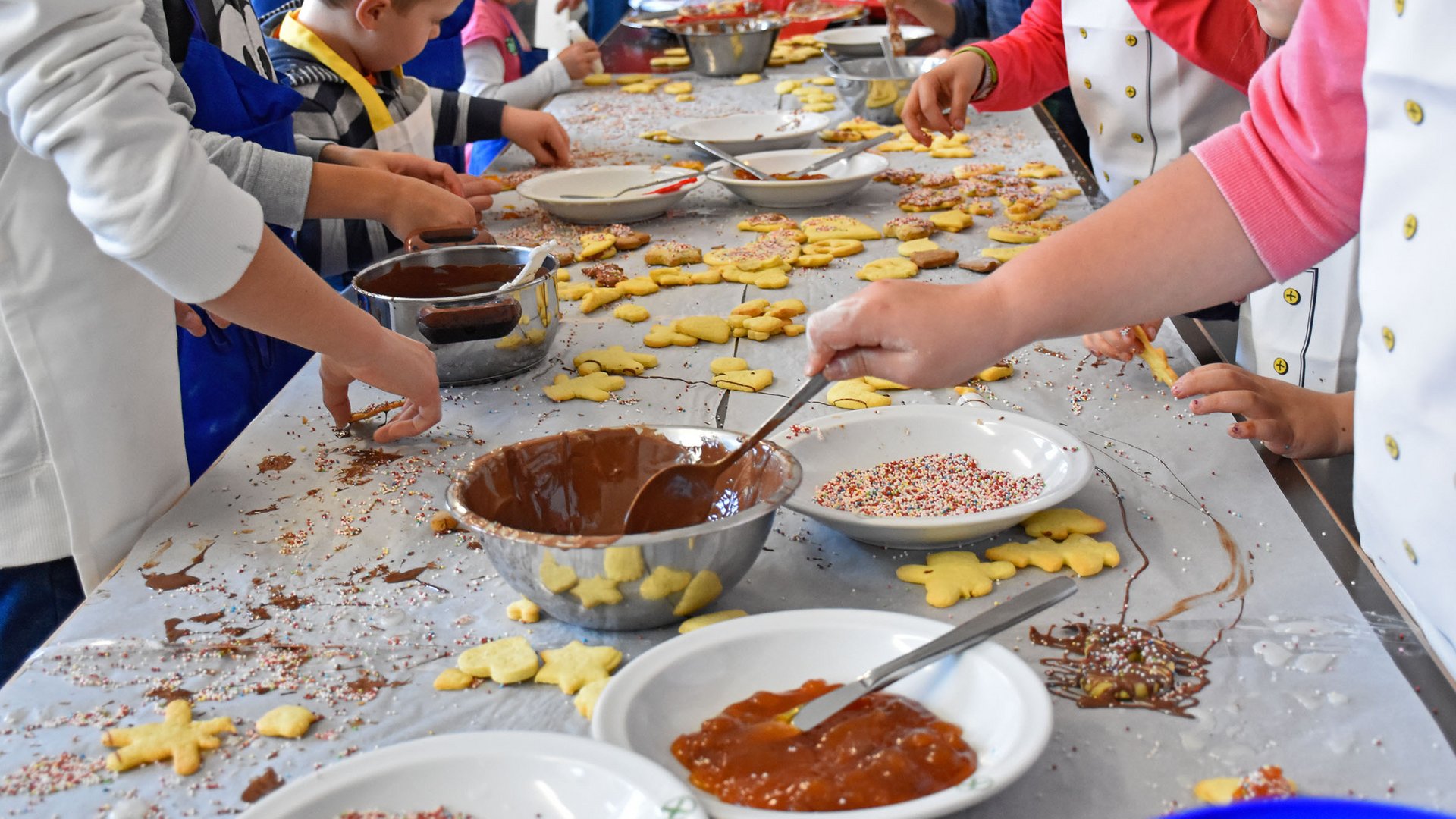 L’Avvento di Terento L'immagine mostra dei bambini che decorano biscotti. Sul tavolo ci sono ciotole con cioccolato fuso, marmellata e zuccherini colorati, mentre i bambini spalmando e decorando i biscotti. Il tavolo è coperto da molti biscotti già pronti e ingredienti.