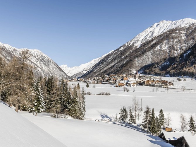 Le località dell’area vacanze Rio Pusteria L'immagine mostra un paesaggio invernale con un villaggio in una valle, circondato da montagne innevate. In primo piano si vedono alberi e colline coperte di neve, mentre il cielo è limpido e blu.
