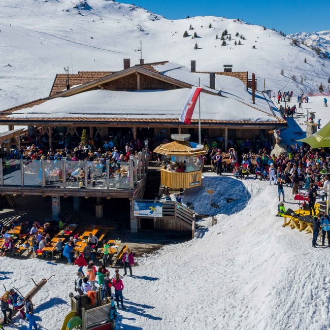Bergrestaurant Jochtal L'immagine mostra un vivace rifugio sciistico con molti ospiti sulla terrazza, circondato da pendii coperti di neve e sciatori, in un paesaggio invernale alpino soleggiato.