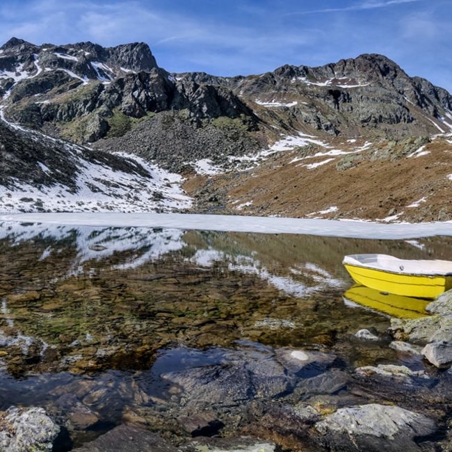 Tiefrastenhütte L'immagine mostra un lago di montagna limpido, parzialmente circondato da neve e ghiaccio, nel mezzo di un paesaggio montuoso roccioso. Una barca a remi gialla si trova sulla riva e le cime innevate si riflettono nelle acque tranquille del lago.