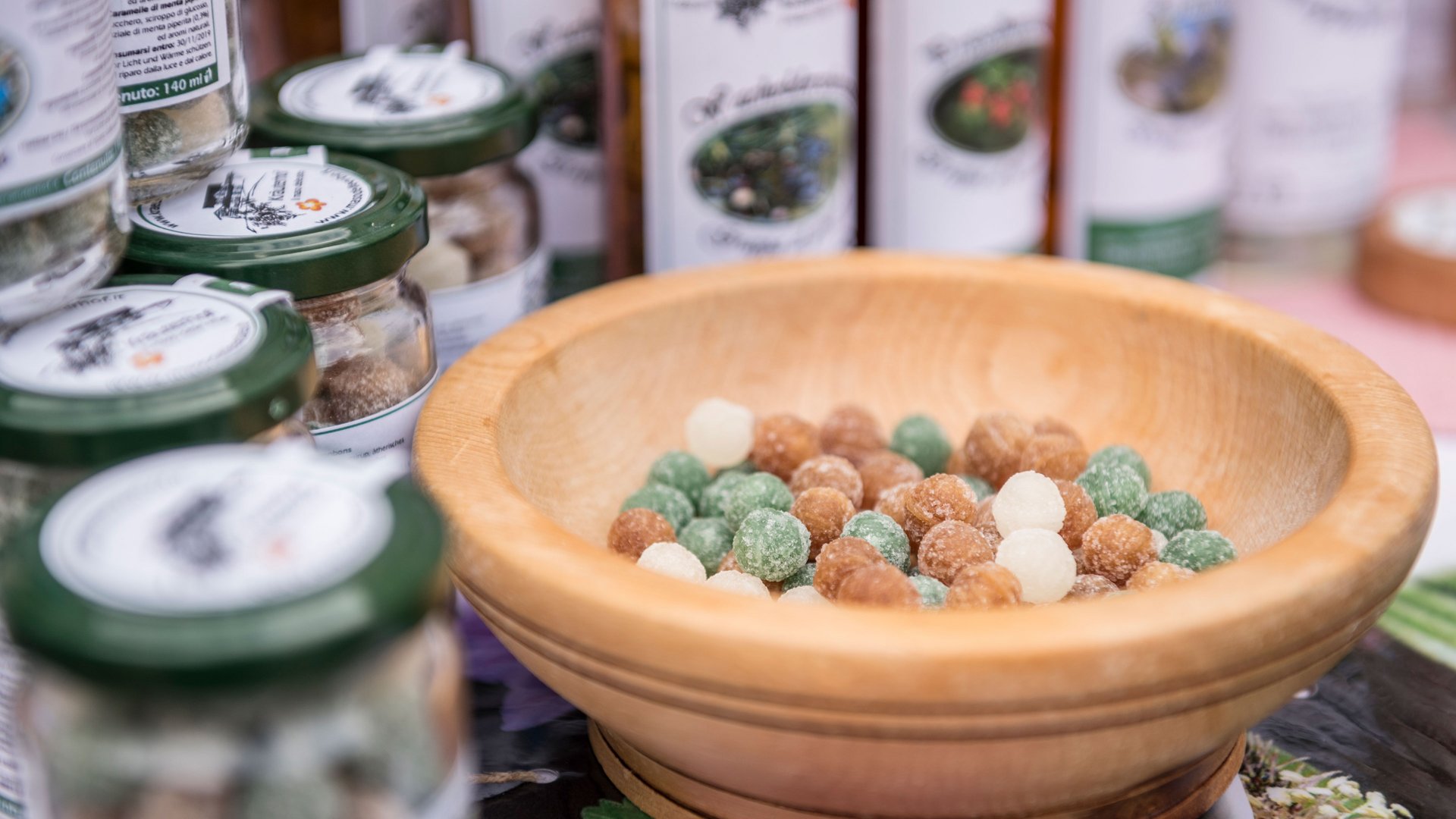 Shopping a Rio di Pusteria (BZ) L'immagine mostra una ciotola di legno piena di caramelle colorate. Sullo sfondo ci sono diversi barattoli con coperchi verdi, anch'essi pieni di caramelle o dolci simili. Sembra un banco di mercato che offre prodotti fatti a mano.