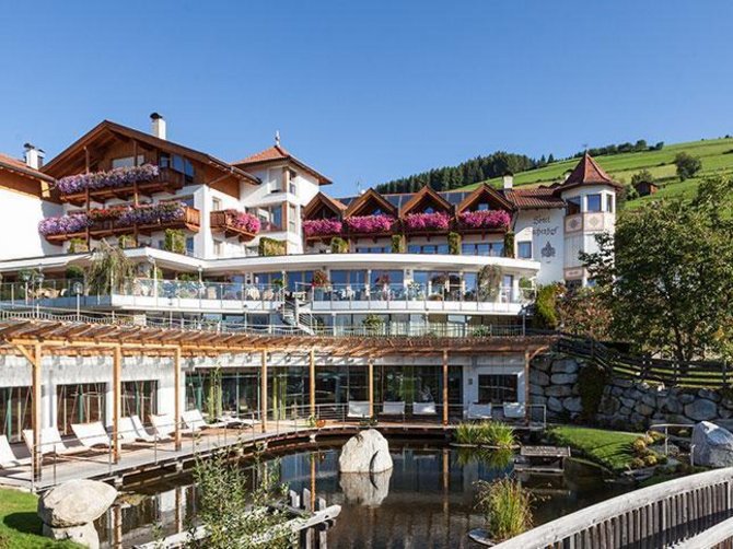 Benvenuti nell’Area Vacanze Sci e Malghe Rio Pusteria! L'immagine mostra un grande ed elegante hotel con più piani e balconi decorati con fiori colorati. In primo piano c'è un piccolo stagno con un ponte, e sullo sfondo si estendono colline verdi e prati.