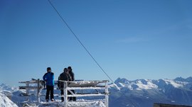 Valles - Jochtal - Steinermandl L'immagine mostra diverse persone con abbigliamento invernale su una piattaforma coperta di neve. Sullo sfondo si vedono montagne innevate sotto un cielo azzurro brillante.